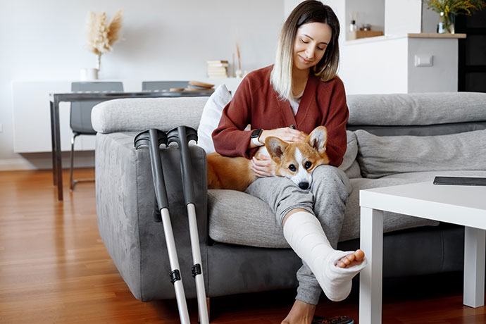Adult woman in her late twenties on couch at home with crutches and orthopedic plaster caress the dog. Fracture of the leg or foot. Concept of rehabilitation and healing. Orthopedics and Traumatology.