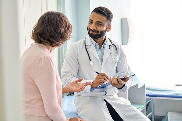 Indian male doctor consulting senior old patient filling form at consultation. Professional physician wearing white coat talking to mature woman signing medical paper at appointment visit in clinic.