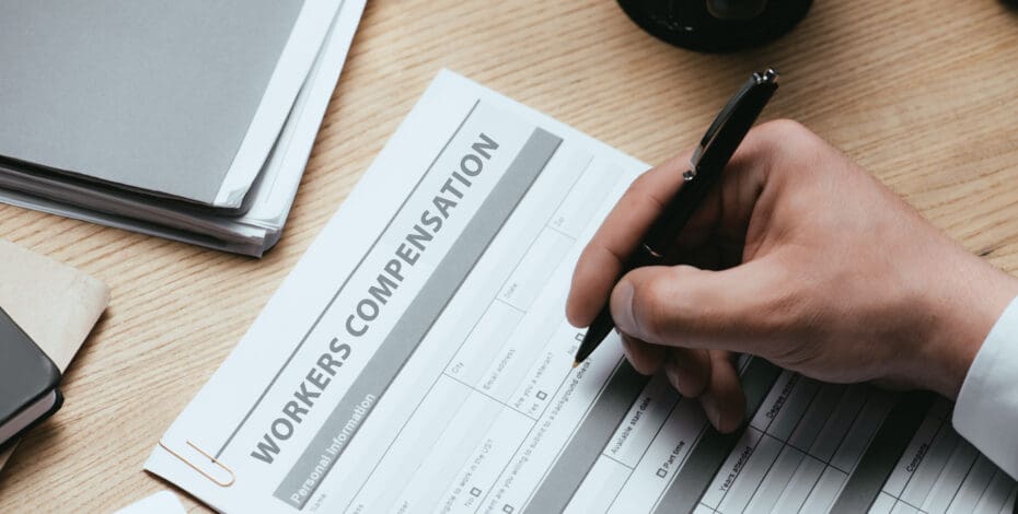 A man's hand holds a pen as he starts to fill out a form labeled 'Workers' Compensation'