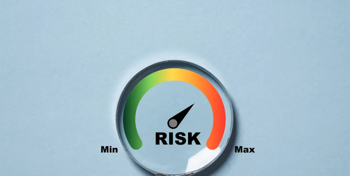 A person holds a magnifying glass. Inside the glass is a scale. The left side of the scale is green with the words 'Min' and the right side is red with the word 'Max.' At the center of the scale is the word 'Risk.'