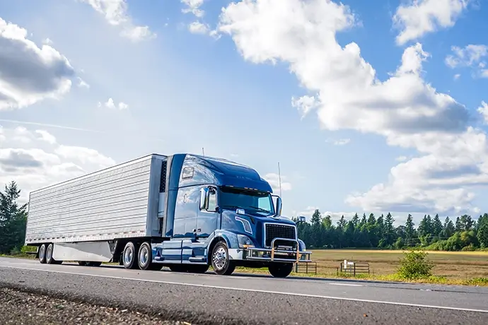 Blue big rig semi truck with grille guard trabsporting frozen cargo in refrigerator semi trailer with skirt spoiler running on the straight road